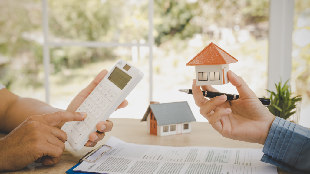 close-up of two hands, one holding a calculator and one holding a miniature model of a house with a desk in the background